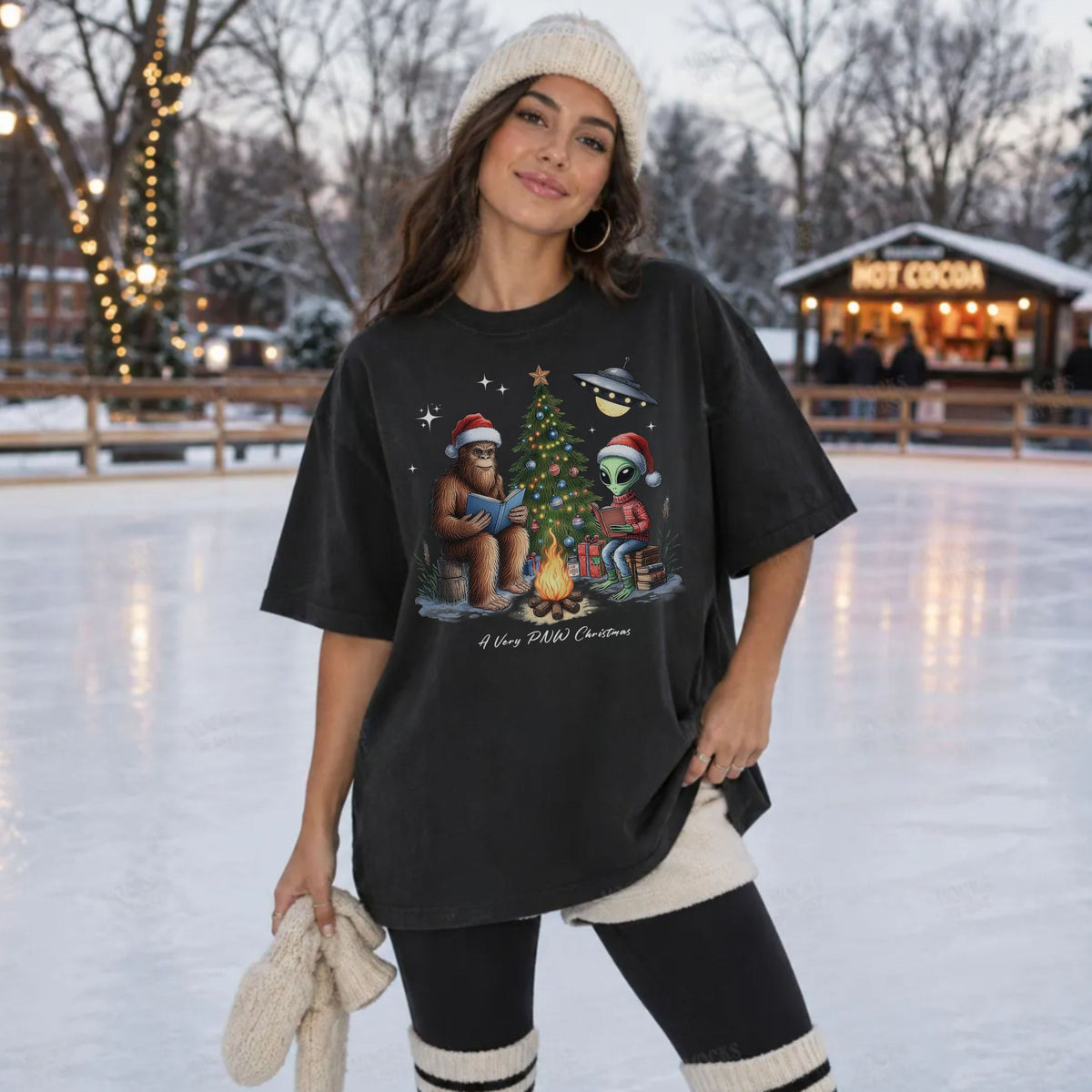 A woman stands on an outdoor ice rink wearing an oversized black graphic T-shirt with a Christmas cartoon design, black leggings, and a cream knit beanie. The background shows snow, trees, string lights, and a hot cocoa shop.