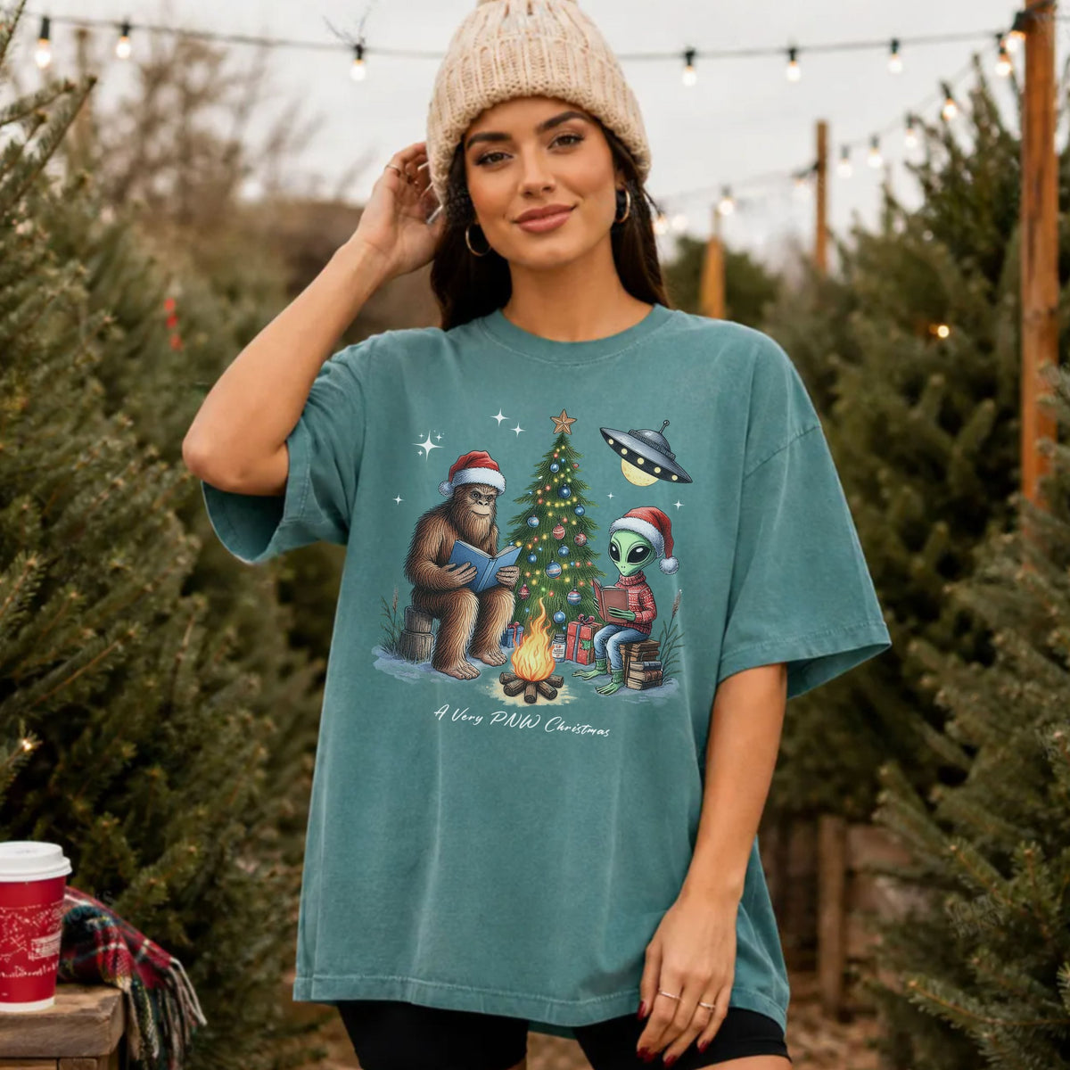 A woman wearing an oversized teal graphic t-shirt with a 'A Very PNW Christmas' cartoon design, standing in a Christmas tree lot.