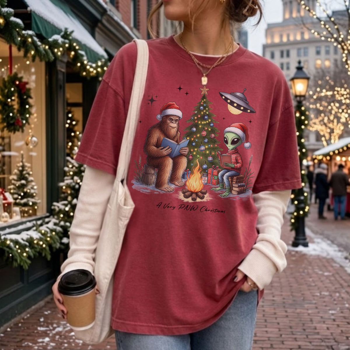 A woman wearing an oversized dusty rose graphic t-shirt with a Christmas bigfoot and alien design, layered over a cream long-sleeve shirt, in an outdoor holiday street setting.