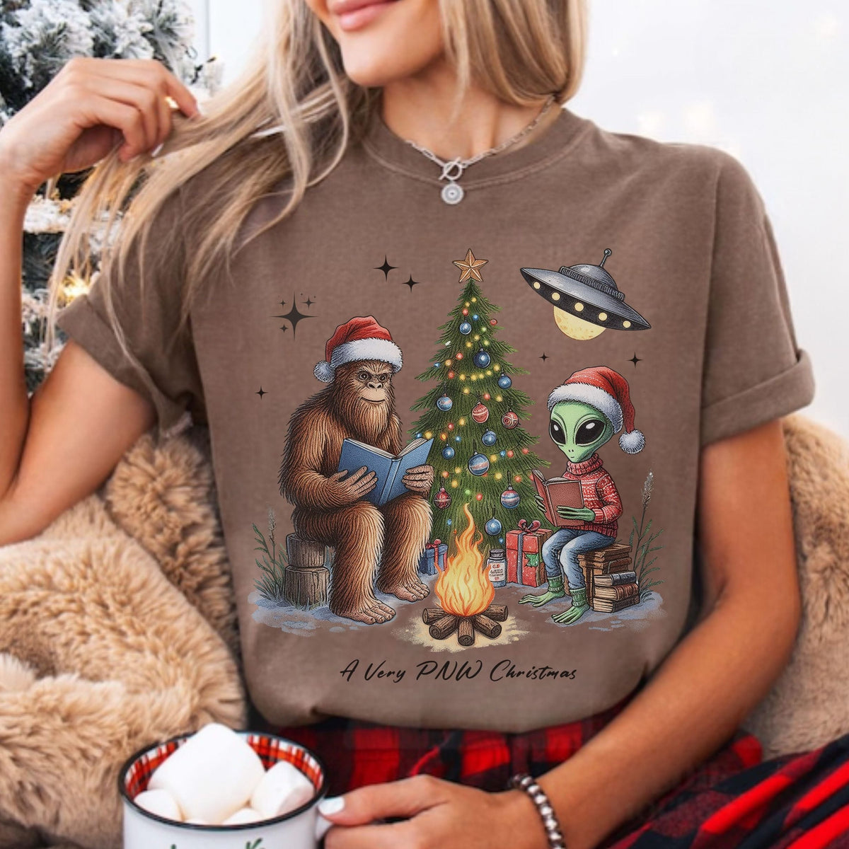 A woman wearing a brown graphic t-shirt with a Christmas-themed design featuring a Bigfoot and an alien sitting by a campfire next to a decorated tree. The text on the shirt reads 'A Very PNW Christmas'.