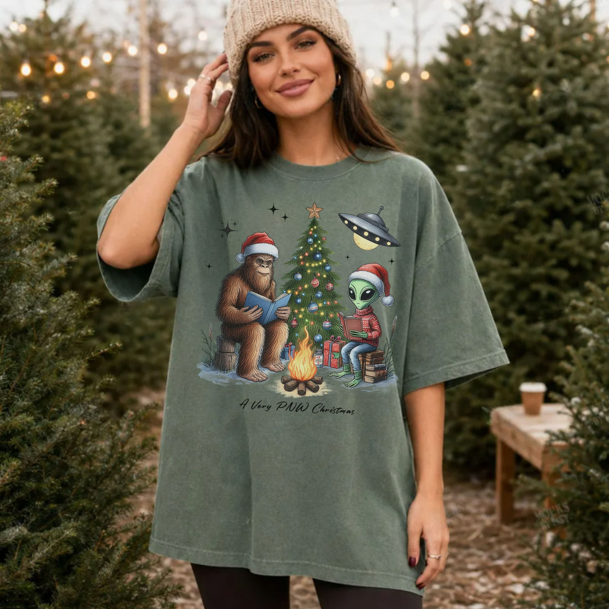 A woman wearing an oversized sage green graphic t-shirt in a Christmas-themed outdoor setting, likely a Christmas tree farm. The t-shirt features a cartoon illustration of a Bigfoot and an alien reading by a Christmas tree with a UFO and gifts.
