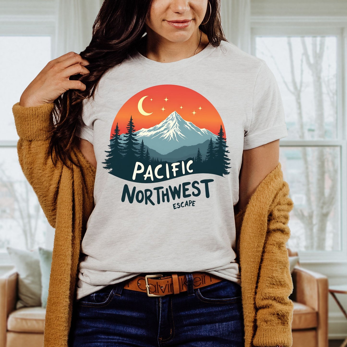 A woman wearing a light gray t-shirt with a colorful 'Pacific Northwest Escape' mountain and sunset graphic, styled with a brown cardigan and jeans in a cozy living room setting.