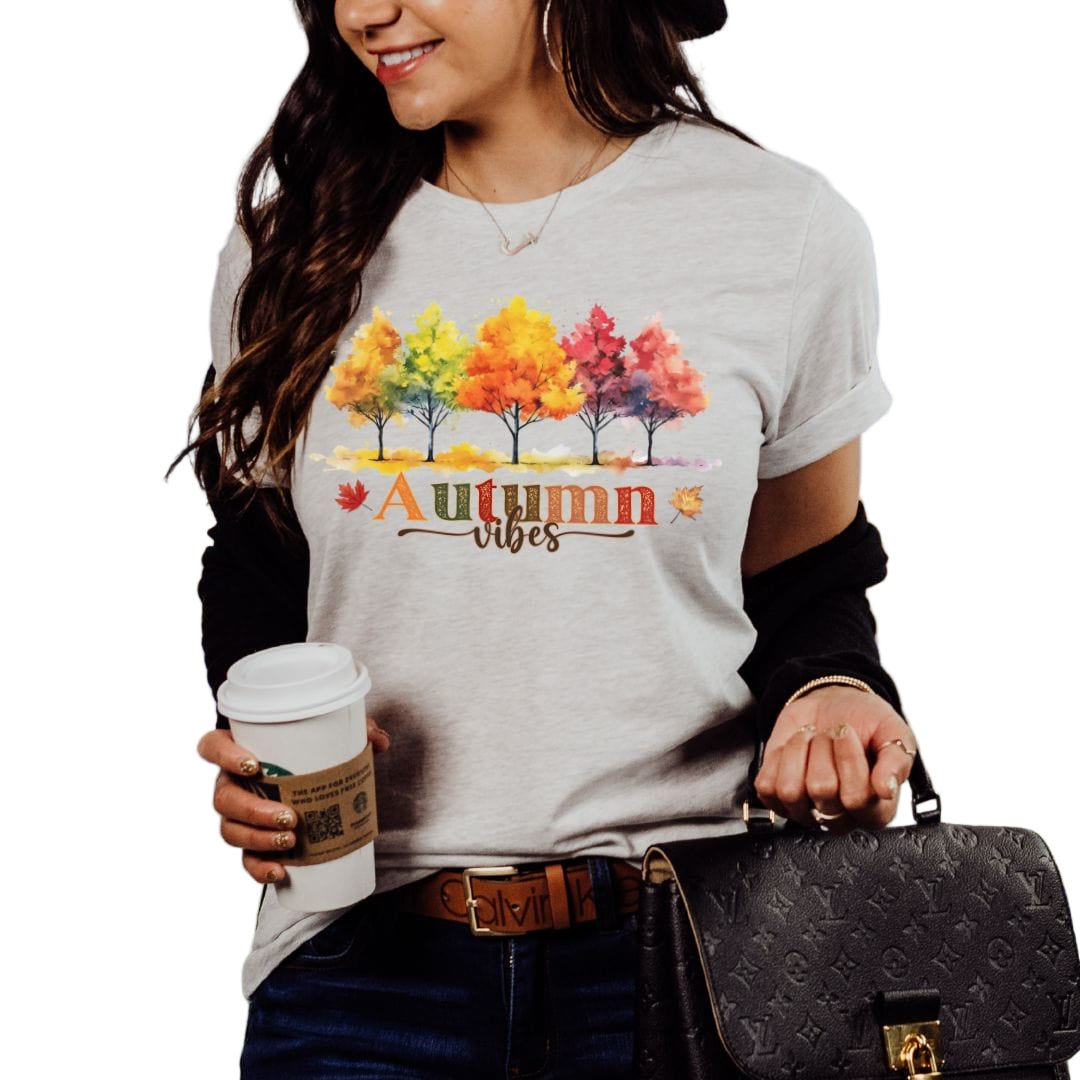 A woman wearing a gray t-shirt with a colorful autumn tree design and the text 'AUTUMN VIBES' on it, holding a coffee cup, and carrying a purse.