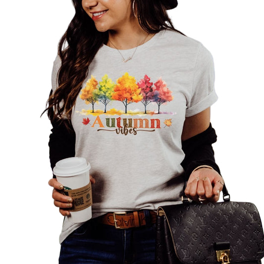 A woman wearing a gray t-shirt with a colorful autumn tree design and the text 'AUTUMN VIBES' on it, holding a coffee cup, and carrying a purse.