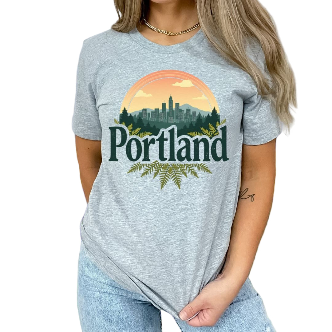 A female model wearing a gray t-shirt with 'Portland' text and a graphic design featuring a cityscape and floral elements.