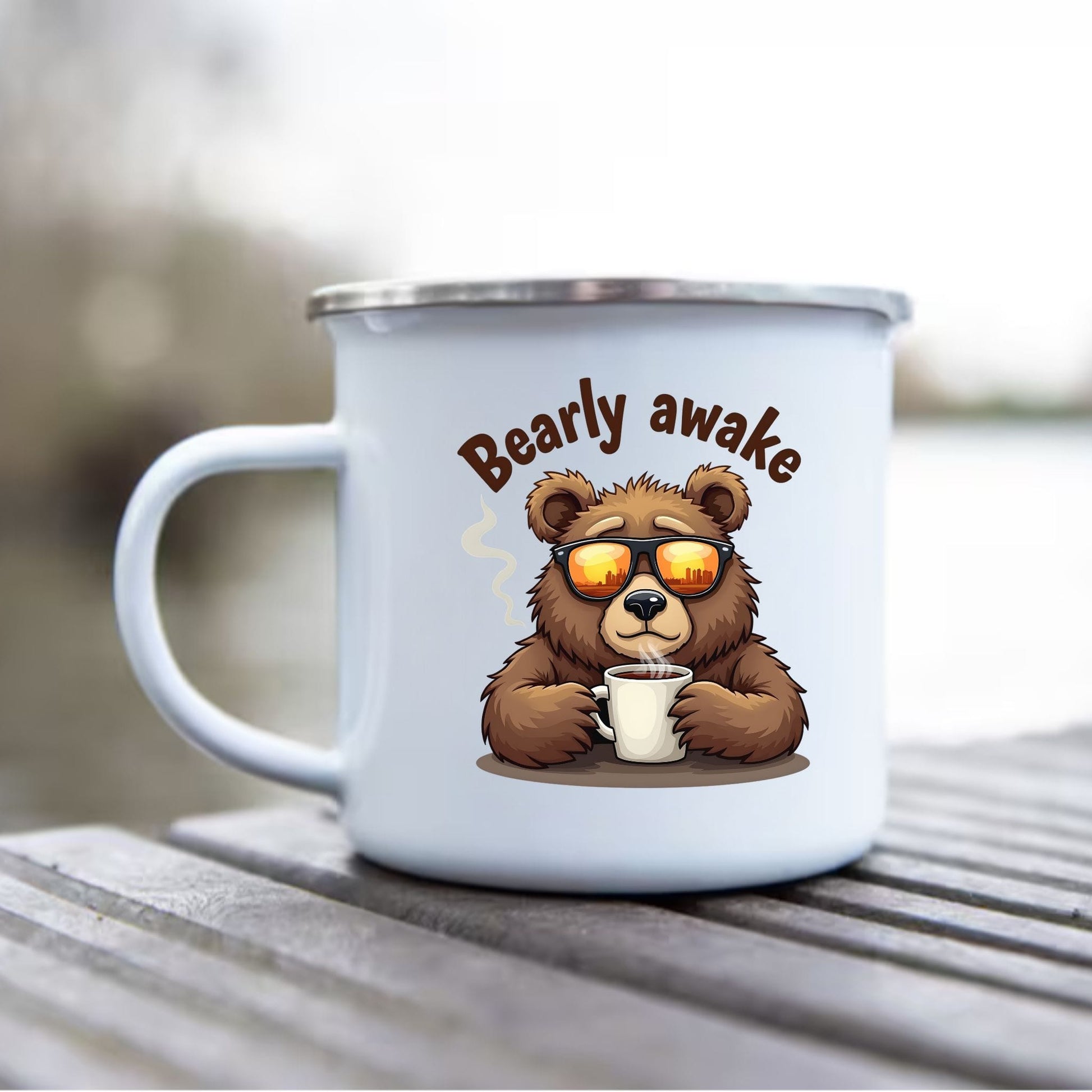A white enamel camping mug with a silver rim sits on a wooden deck. The mug features a cartoon graphic of a brown bear wearing orange-tinted sunglasses and holding a coffee cup, with the text 'Bearly awake' above it.