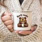 A person in a chunky cream knit sweater holds a white enamel-style mug with a cartoon brown bear wearing orange-tinted sunglasses and the words “Bearly awake” above it.
