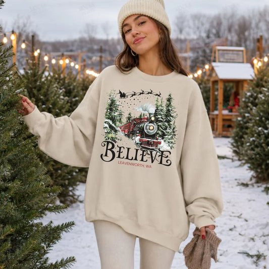A woman wearing a cream-colored crewneck sweatshirt with a festive Christmas graphic of a train and the word 'BELIEVE' stands in a snowy Christmas tree lot.