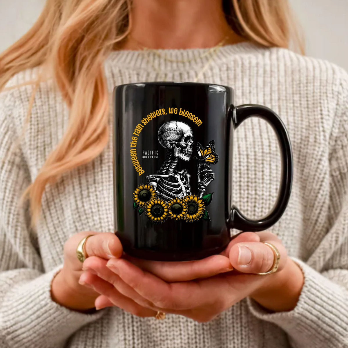 A person in a beige ribbed sweater holds a black ceramic mug with a printed design of a skeleton, sunflowers, and a butterfly. The mug features the text 'Pacific Northwest' in yellow and black.