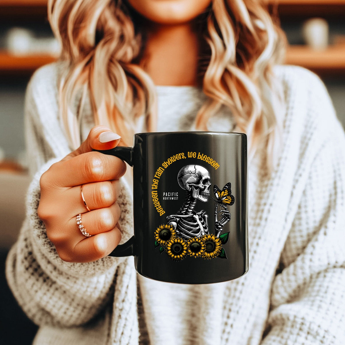 A woman in a white knit sweater holds a black ceramic mug with a graphic design. The design features a white skeleton, yellow sunflowers, a butterfly, and gold text that reads 'Between the rain showers, we bloom'.