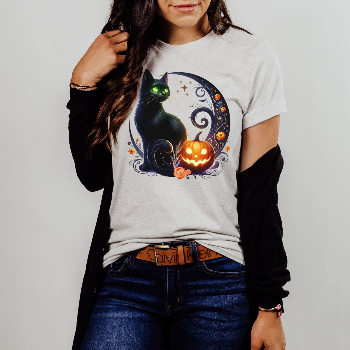 A woman wearing a light gray heather t-shirt with a Halloween graphic of a black cat and a jack-o'-lantern, styled with a black cardigan, blue jeans, and a brown belt.