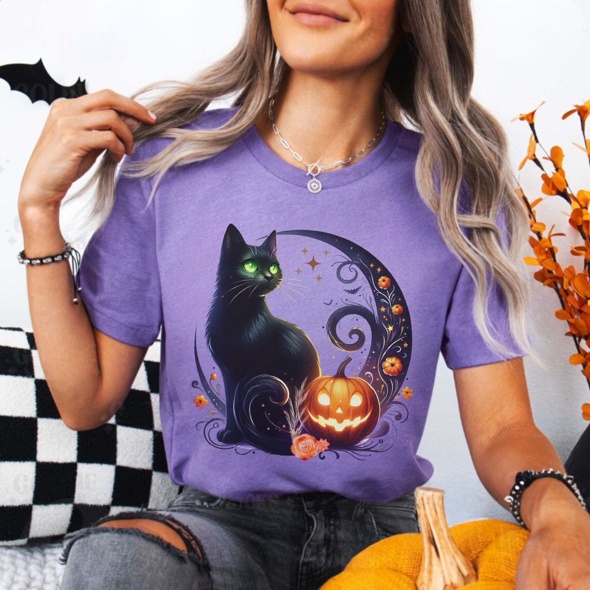 A woman wearing a purple short-sleeve graphic t-shirt with a black cat and jack-o'-lantern design, paired with ripped gray jeans, sitting in a cozy indoor setting.