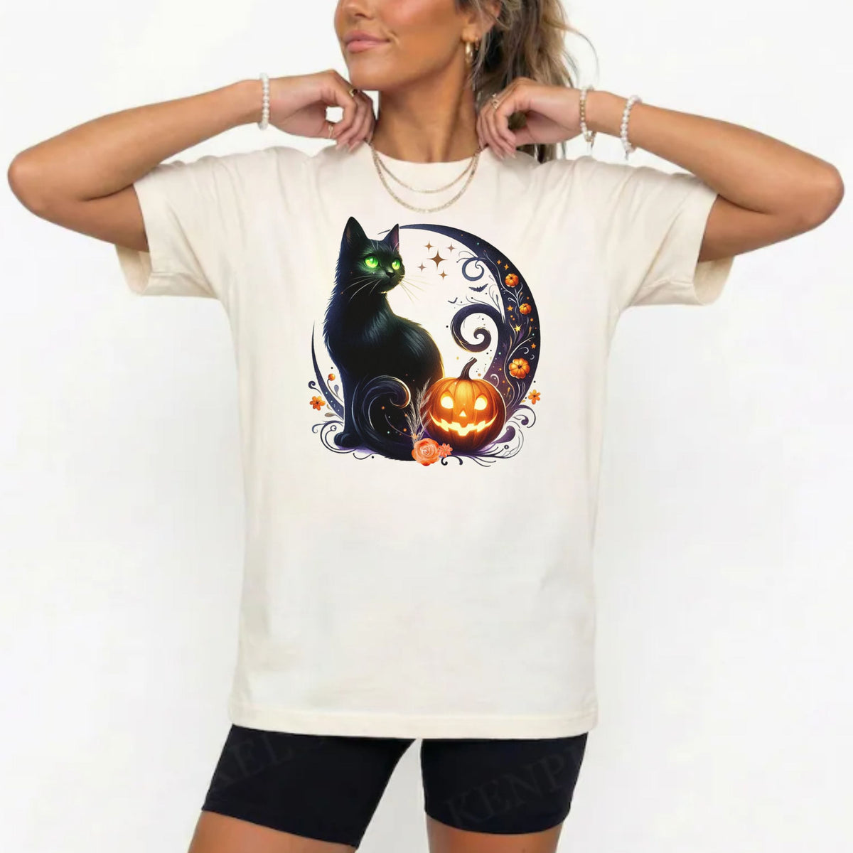 A woman wearing a cream-colored t-shirt with a Halloween graphic of a black cat, orange jack-o'-lantern, and a crescent moon, paired with black bike shorts against a white background.