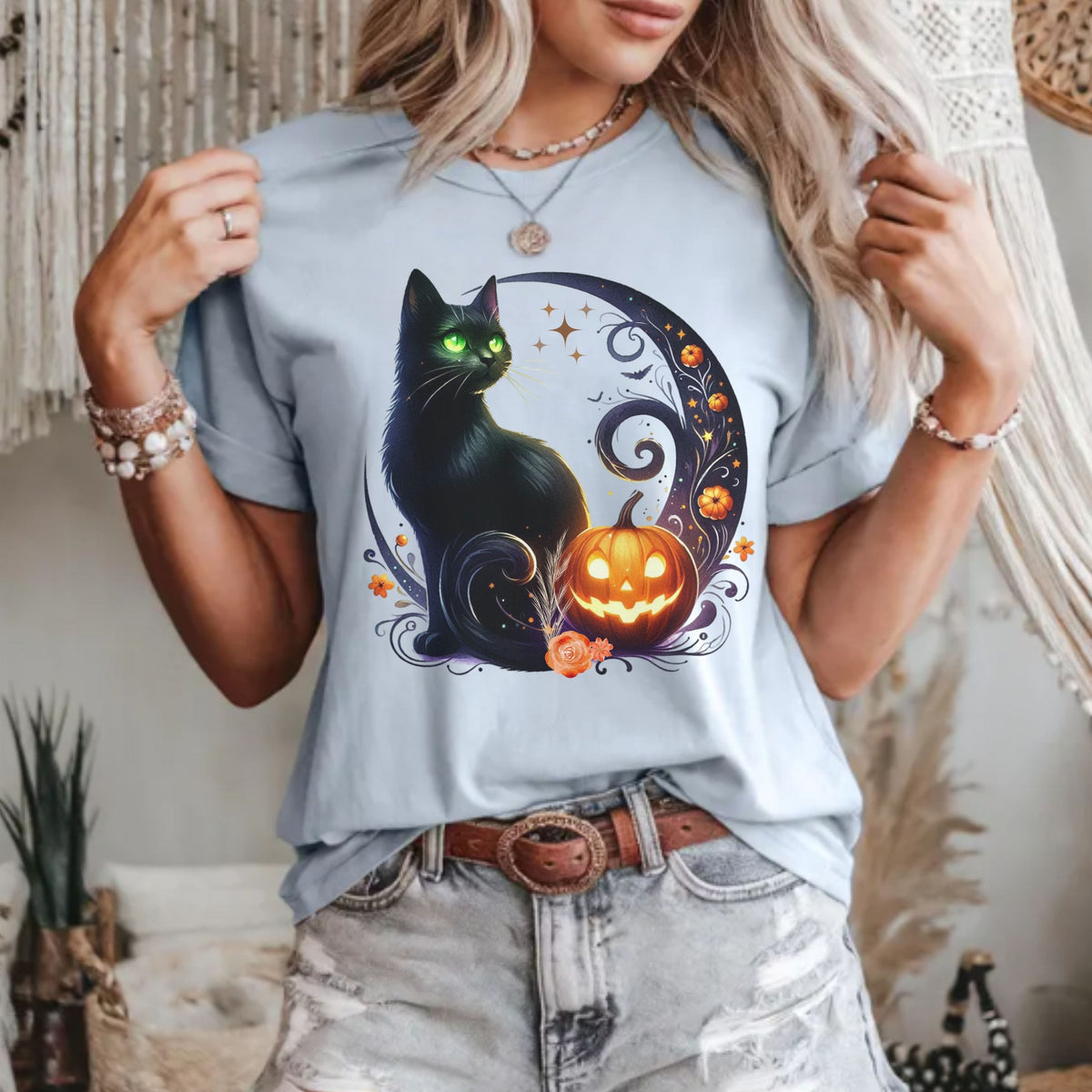 A woman wearing a light blue graphic t-shirt featuring a black cat and a glowing jack-o'-lantern in a crescent moon design, paired with distressed denim shorts.