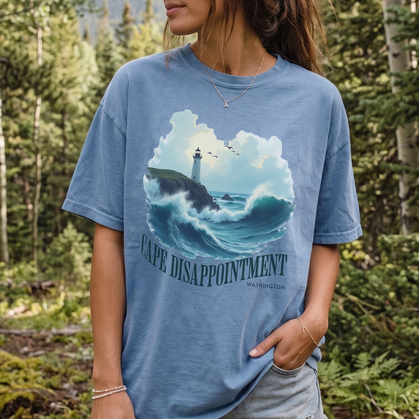 Woman wearing a blue Comfort Colors t-shirt with a Cape Disappointment Washington graphic featuring a coastal lighthouse, crashing ocean waves, and green arched text.