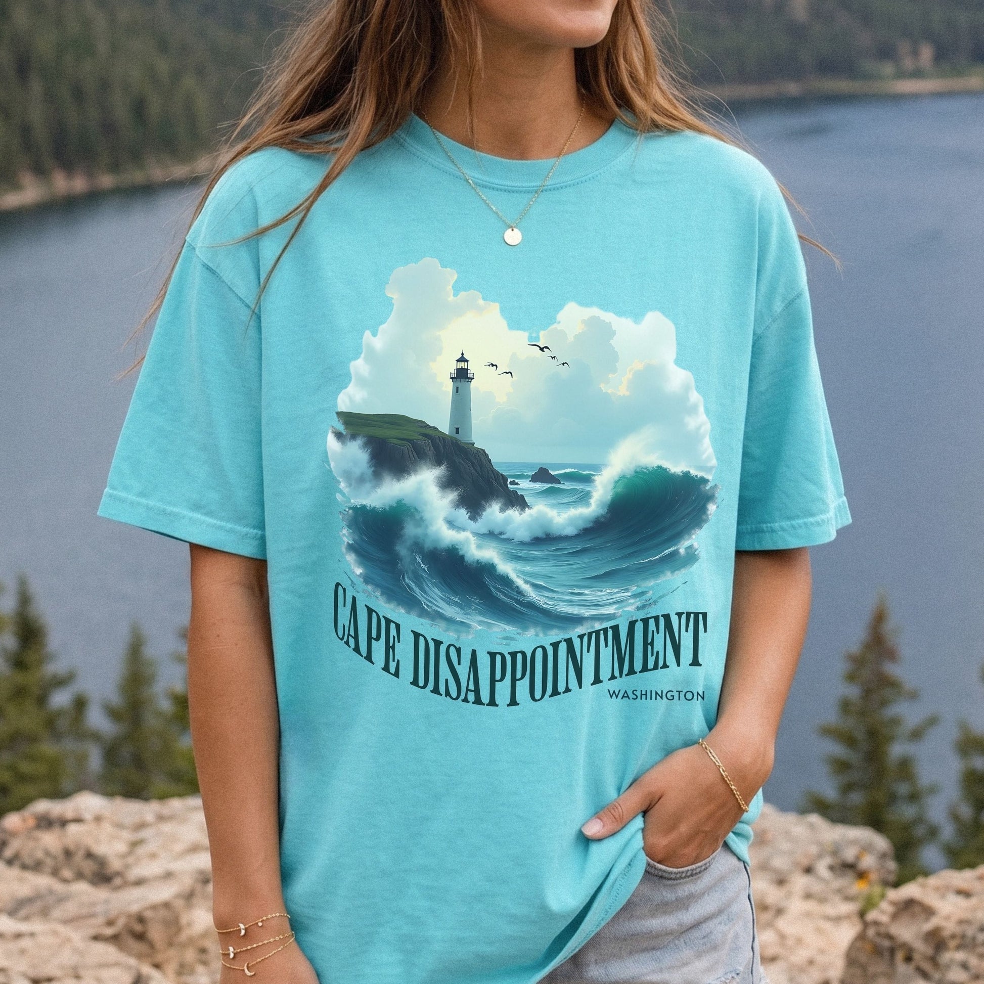 A woman wearing a light blue graphic t-shirt with a lighthouse and crashing waves design, standing outdoors with a lake and forest background.