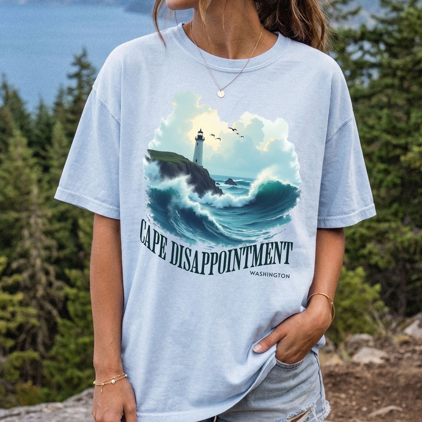 Woman wearing a chambray Comfort Colors t-shirt with a Cape Disappointment Washington graphic featuring a coastal lighthouse, crashing ocean waves, and green arched text.