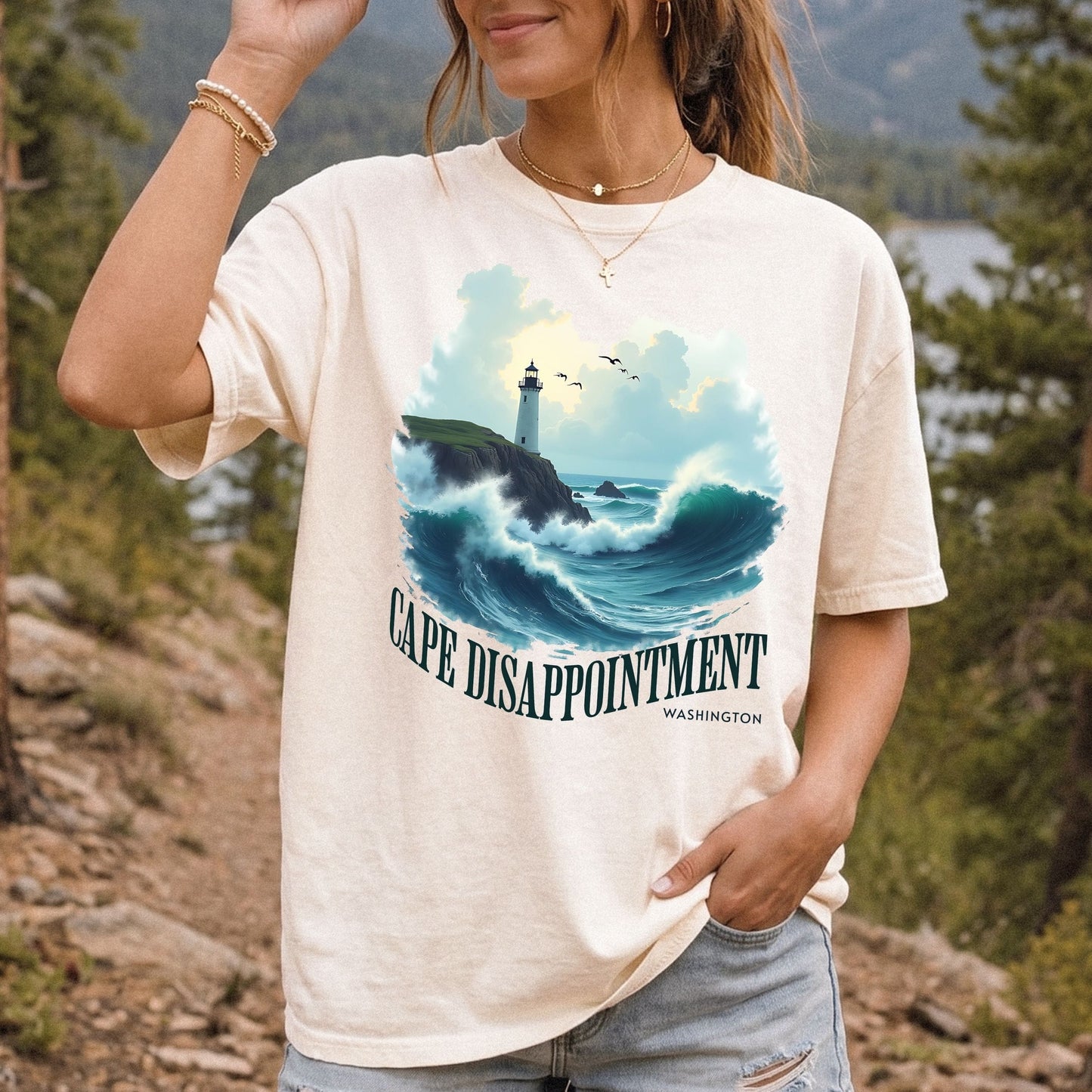 Woman wearing an ivory Comfort Colors t-shirt with a Cape Disappointment Washington graphic featuring a coastal lighthouse, crashing ocean waves, and green arched text.