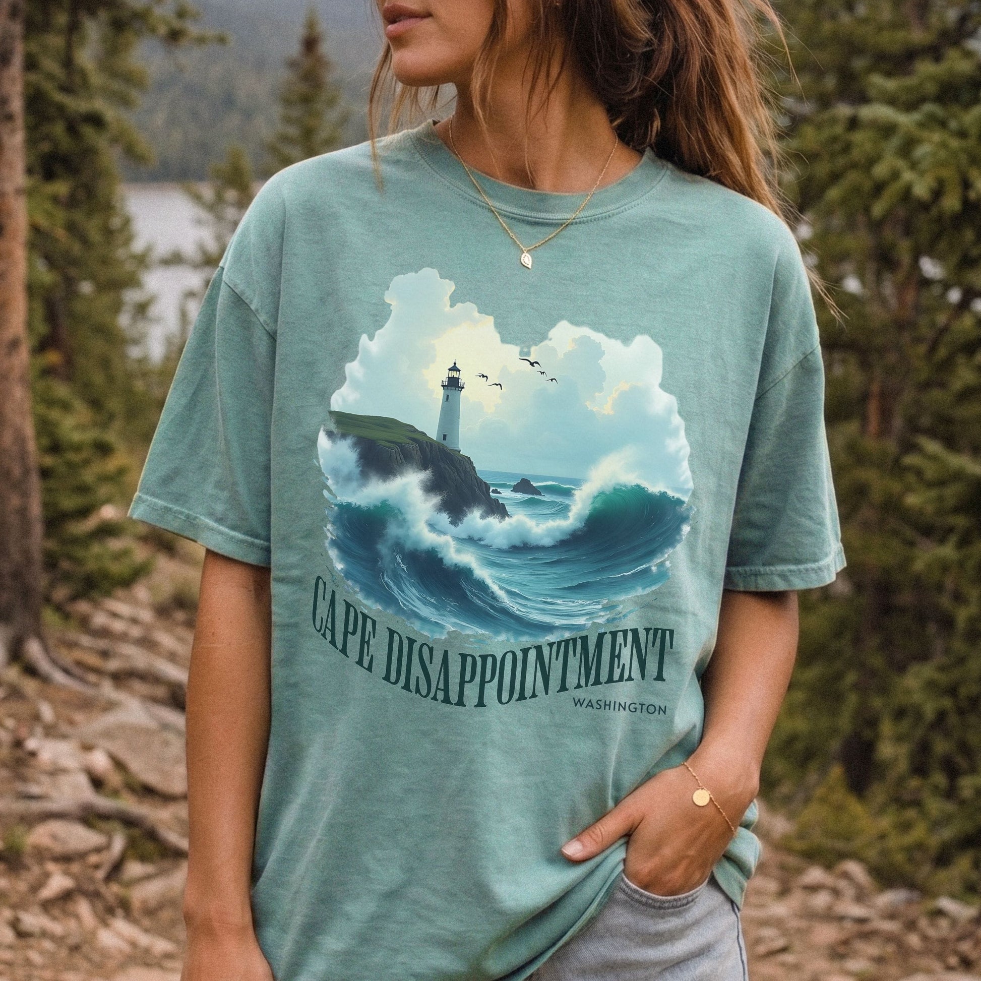 Woman wearing a seafoam green, Comfort Colors t-shirt with a Cape Disappointment Washington graphic featuring a coastal lighthouse, crashing ocean waves, and green arched text.