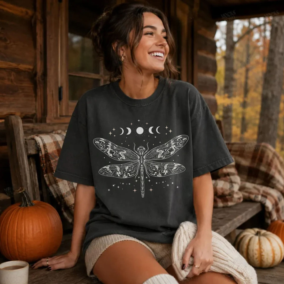 A woman sitting on a rustic wooden porch wearing a dark charcoal t-shirt with a white celestial dragonfly and moon graphic. The background features a cozy fall atmosphere with pumpkins, a plaid blanket, and an autumn forest.