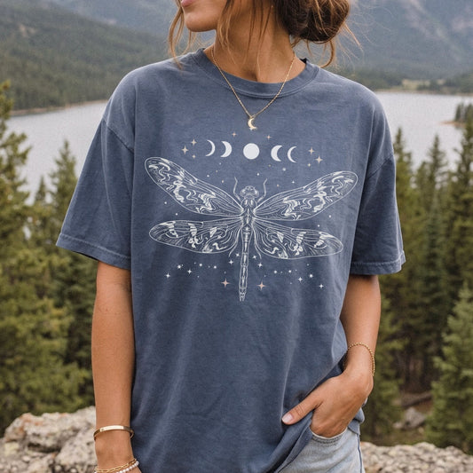 A woman wearing a washed slate blue t-shirt with a white celestial dragonfly and moon graphic, standing outdoors in front of a scenic lake and pine forest background.