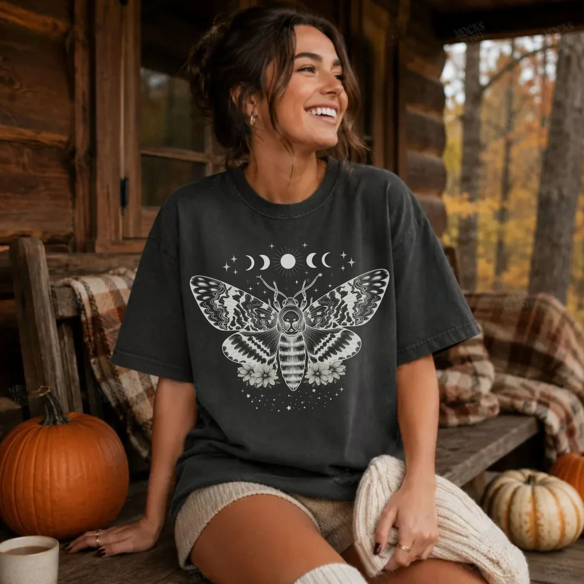 A woman sitting on a rustic wooden porch wearing a dark charcoal gray t-shirt with a white moth and moon graphic. The background features a log cabin wall, autumn trees, and pumpkins.