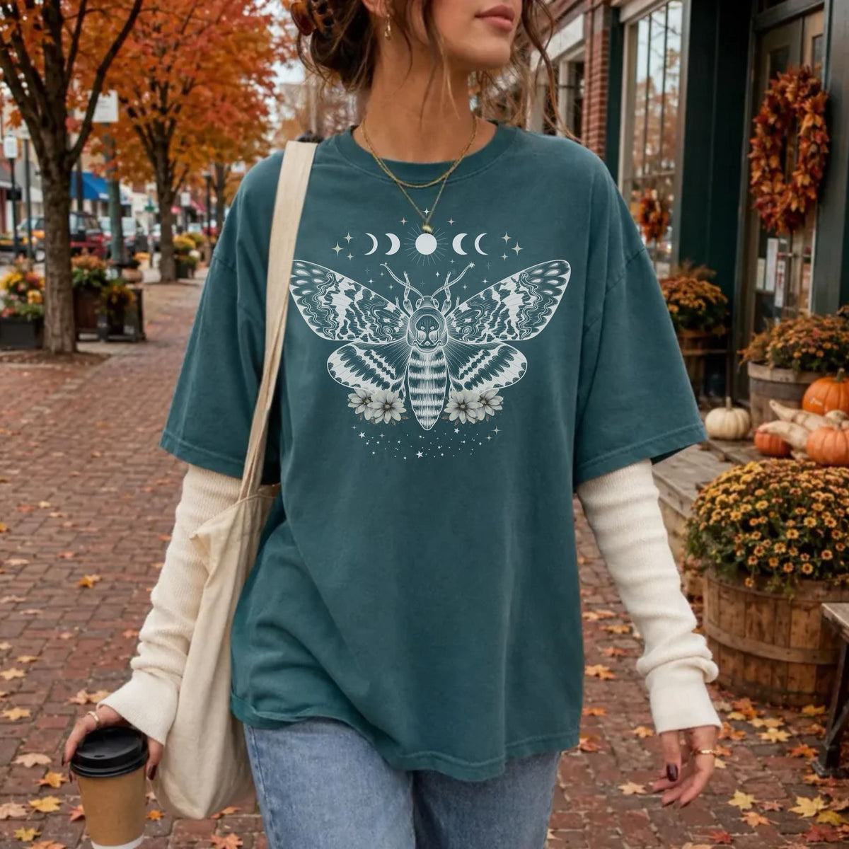 A woman wearing an oversized teal graphic t-shirt with a white moth and moon design, layered over a cream long-sleeve shirt, walking on a brick street with autumn leaves and pumpkins in the background.