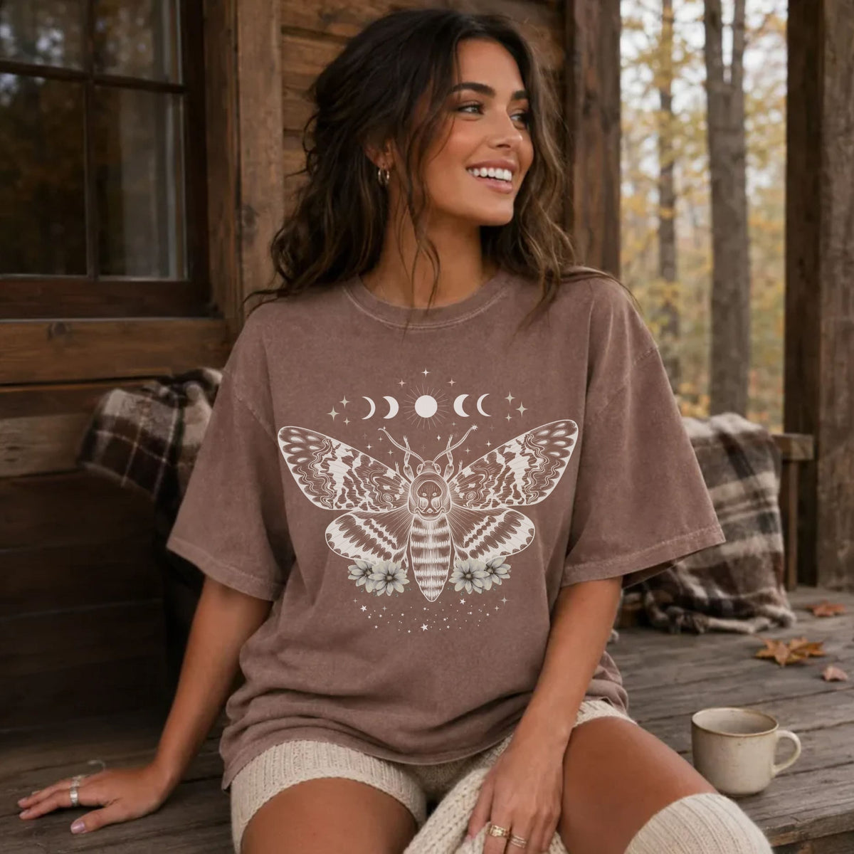 A woman sitting on a wooden porch wearing an oversized brown t-shirt with a white celestial moth and moon graphic, paired with cream knit shorts and a mug nearby.