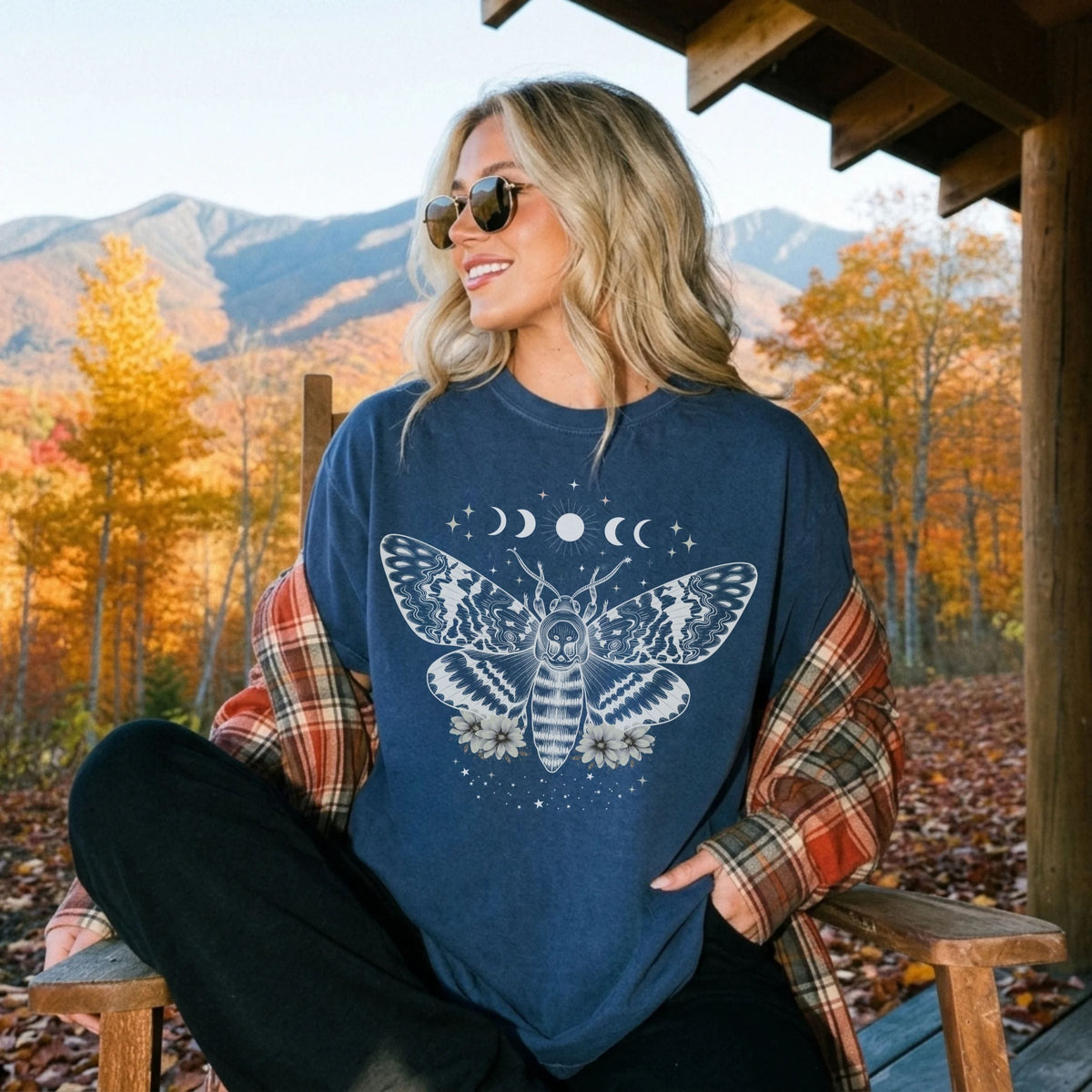 A woman wearing a navy blue crewneck sweatshirt with a white celestial moth and moon graphic, layered over a rust-orange plaid flannel shirt. She is sitting on a wooden porch with a mountain and autumn forest background.