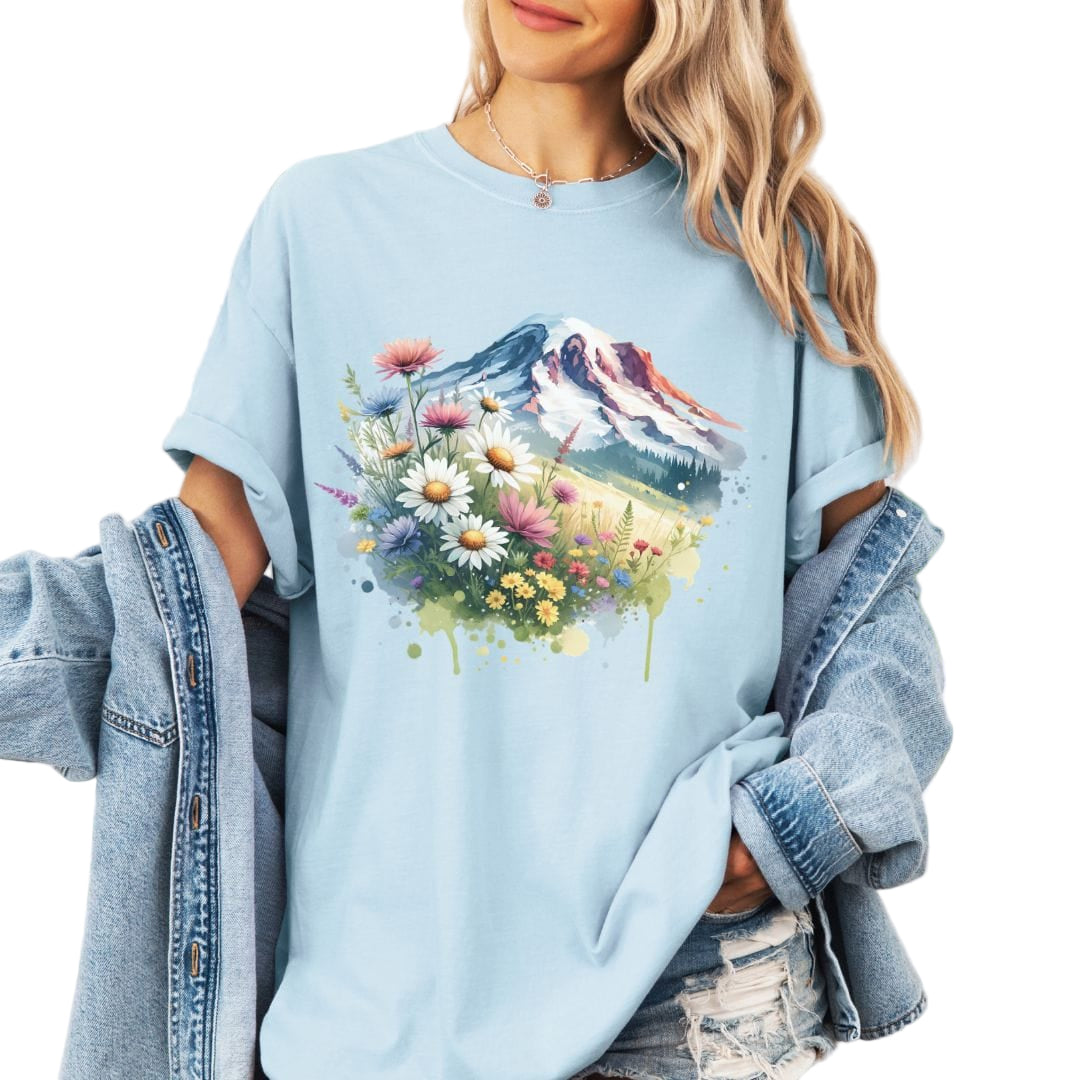 A woman wearing a light blue t-shirt with a vintage-style floral and mountain design, posing outdoors.