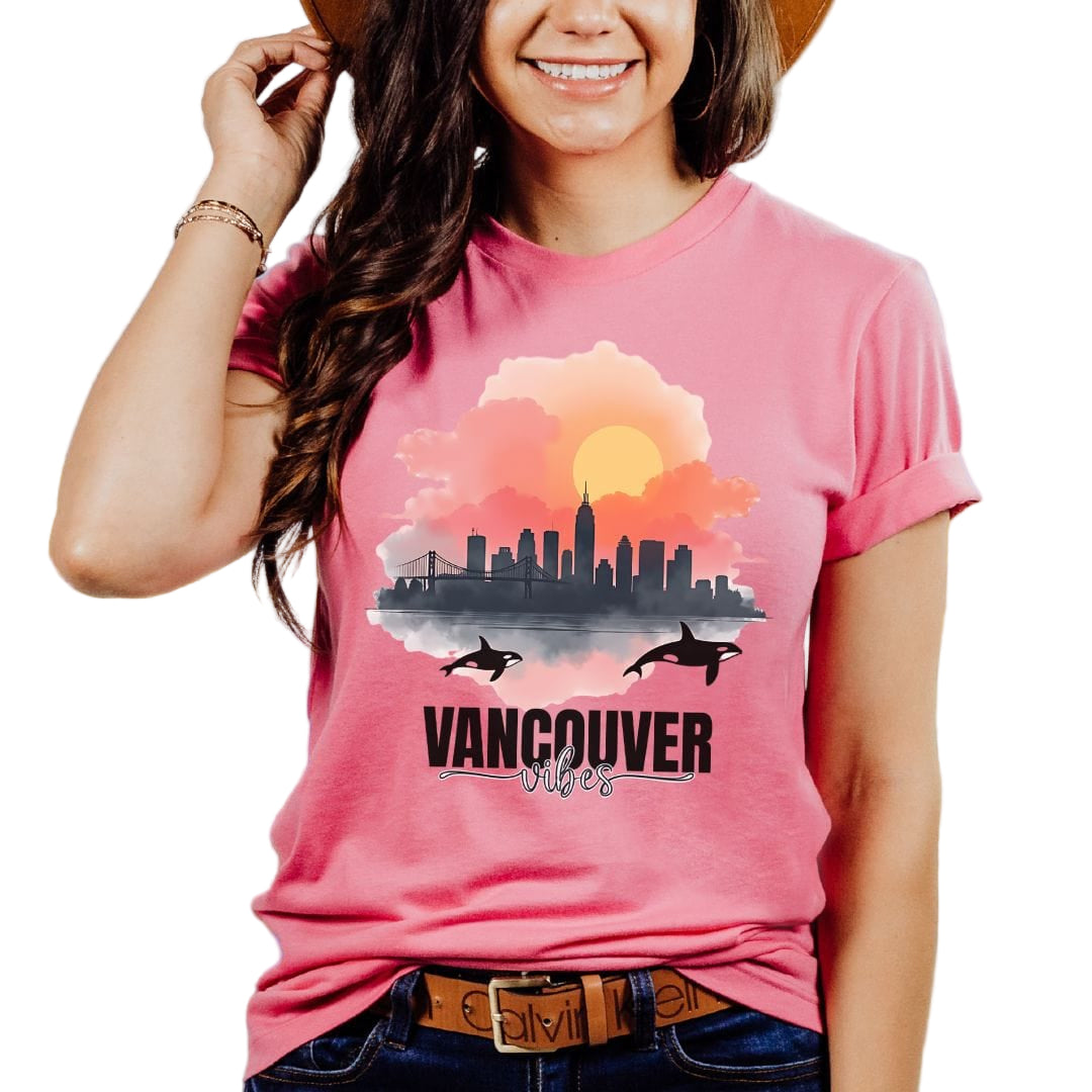 A woman wearing a pink t-shirt with a watercolor style print of Vancouver cityscape and the word 'VANCOUVER' on it, smiling and wearing a wide-brimmed hat.