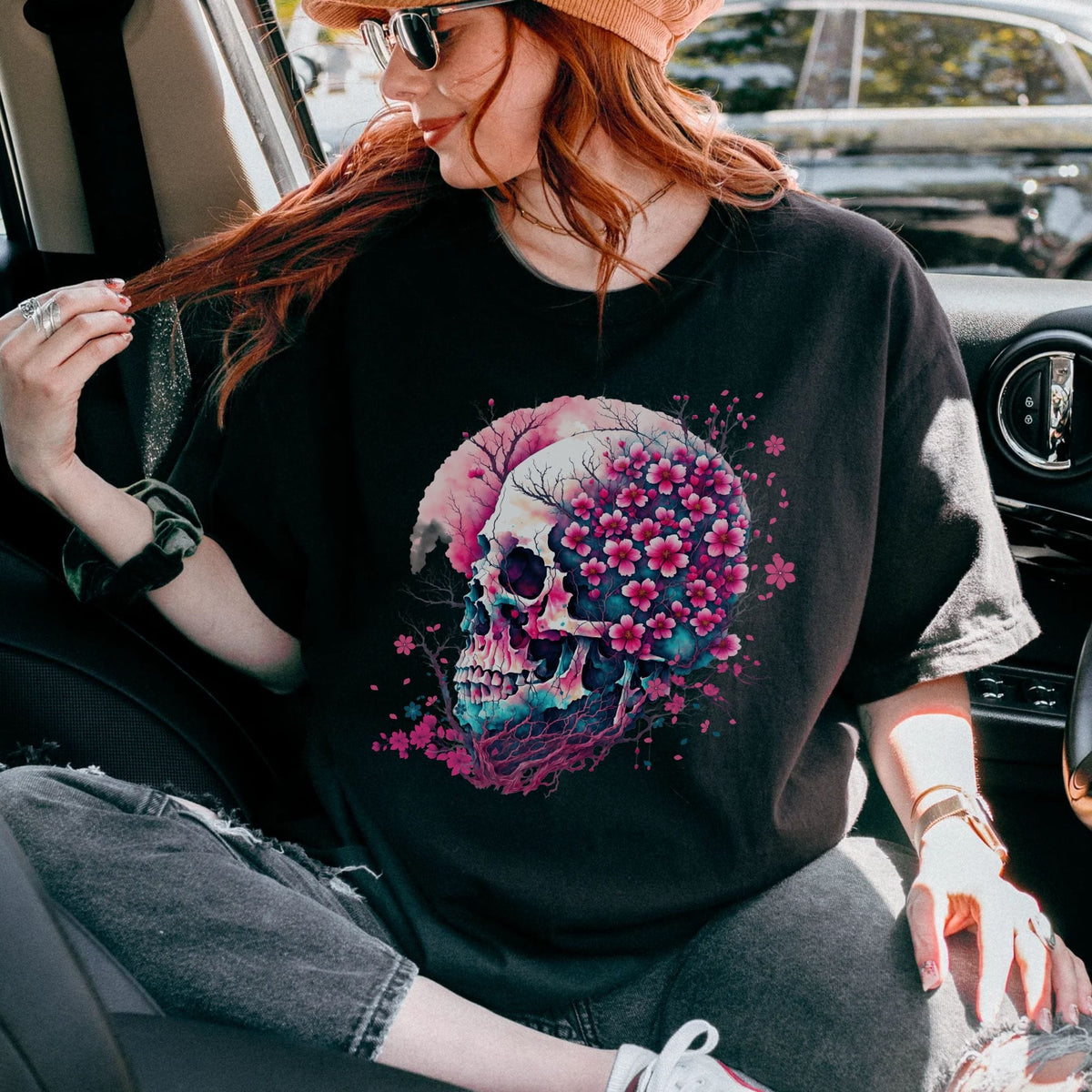 A woman wearing a black graphic t-shirt with a colorful skull and pink cherry blossom design, sitting inside a car.