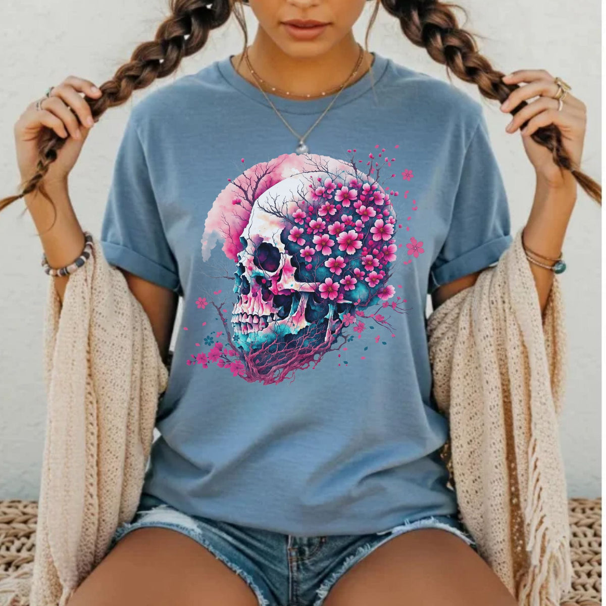 A woman wearing a light blue graphic t-shirt with a skull and pink flowers design, styled with a beige knit cardigan and denim shorts.