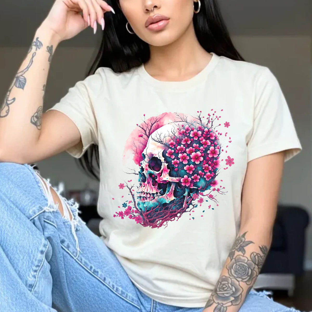 A woman wearing a cream-colored t-shirt with a vibrant graphic of a skull surrounded by pink cherry blossoms and blue foliage, paired with light-wash ripped jeans.