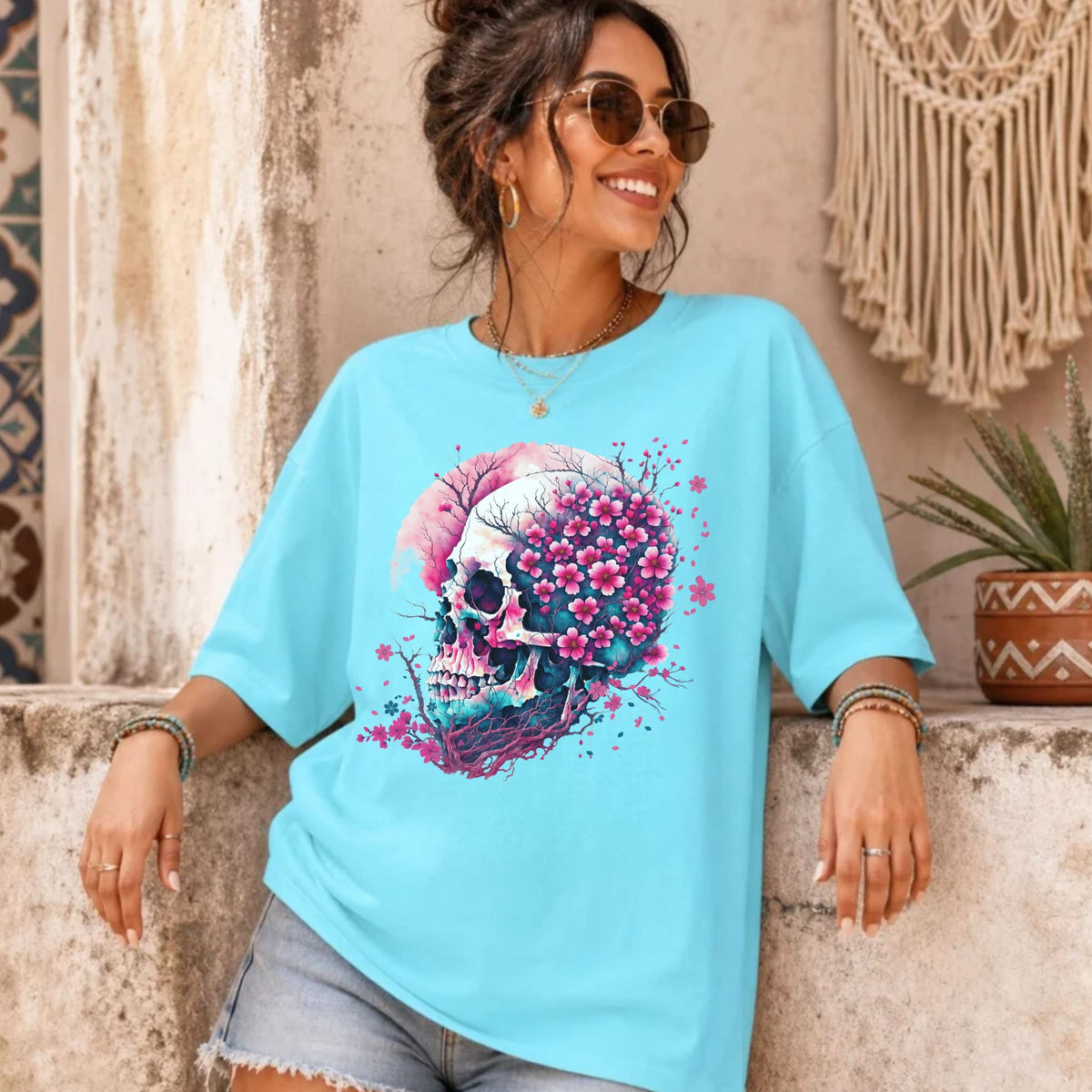 A woman wearing a light blue oversized t-shirt with a colorful skull graphic, styled with denim shorts in a rustic, bohemian-themed setting.