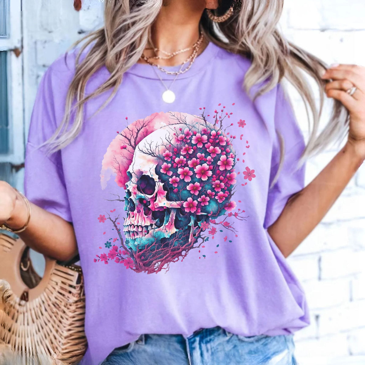 A woman wearing a lavender purple t-shirt with a colorful graphic of a skull filled with pink cherry blossoms, paired with blue jeans and layered gold necklaces.