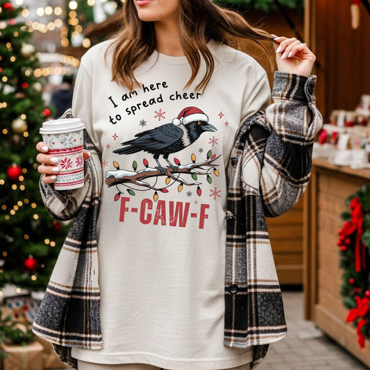 A woman wearing a cream-colored Christmas graphic sweatshirt with a raven and the text 'I am here to spread cheer' and 'F-CAW-F'. She has a black, white, and gray plaid flannel shirt draped off her shoulders and is holding a holiday-themed coffee cup in a festive, decorated indoor setting.