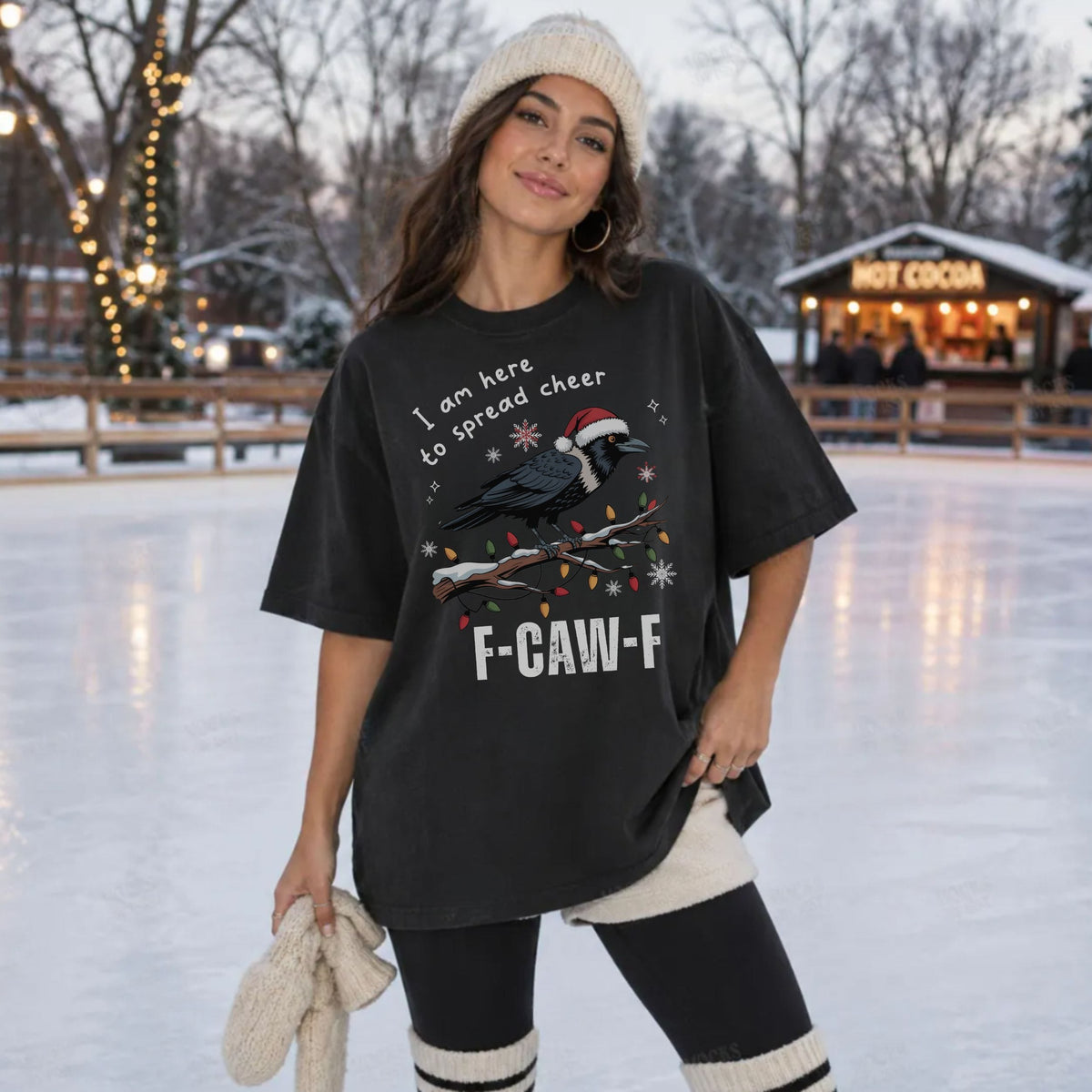 A woman stands on an outdoor ice rink wearing an oversized black graphic T-shirt with a bird and holiday text, black leggings, cream knit socks, and a cream knit beanie. The background shows snow, trees, string lights, and a hot cocoa stand.