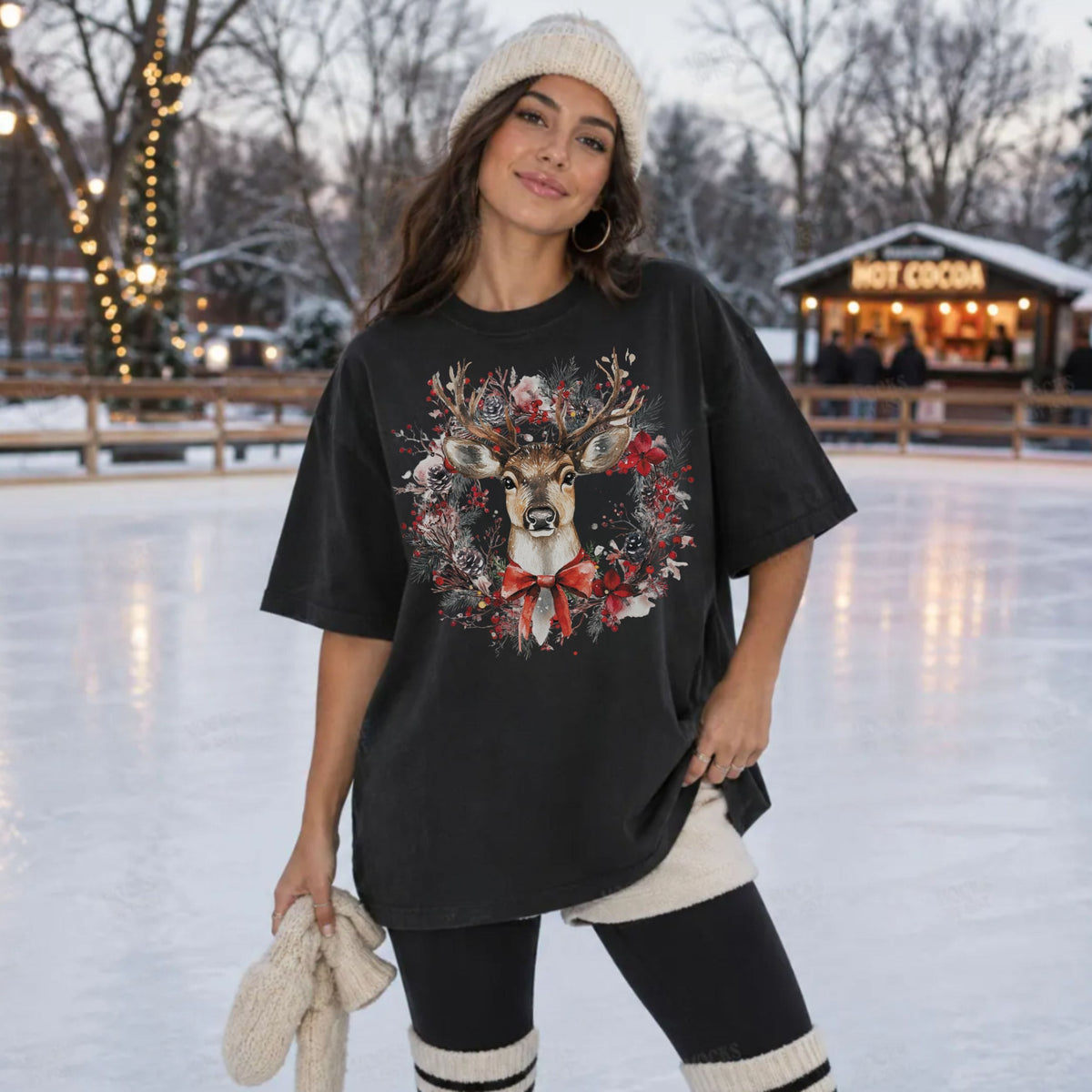 A woman stands on an outdoor ice rink wearing an oversized black graphic T-shirt with a deer and wreath design, black leggings, a cream knit beanie, and cream knit boot covers. A hot cocoa shop is visible in the background.