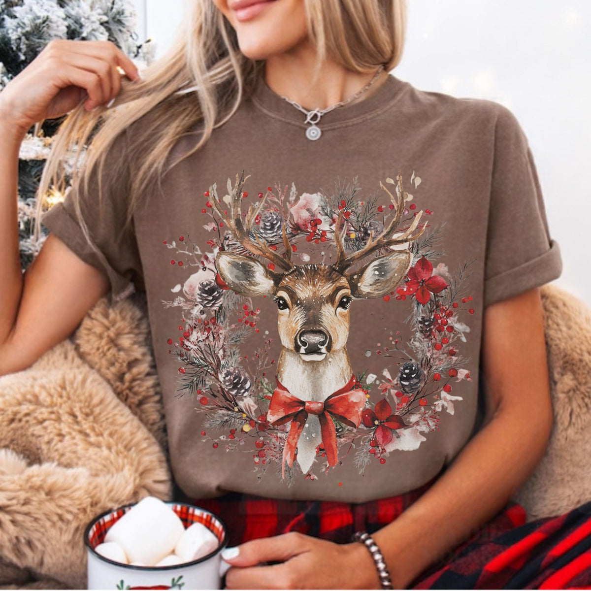 A woman wearing a brown short-sleeve t-shirt with a festive Christmas deer graphic, sitting in a cozy setting with a mug of marshmallows.