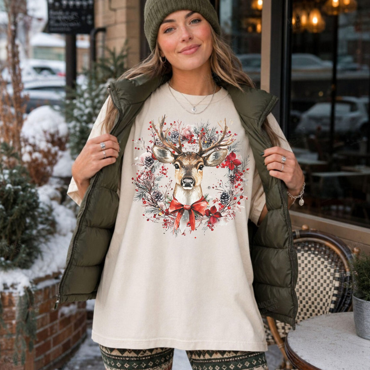 A woman wearing a cream-colored t-shirt with a festive deer and floral wreath graphic, styled with an olive green puffer vest, a green beanie, and patterned leggings, standing outdoors in front of a storefront.