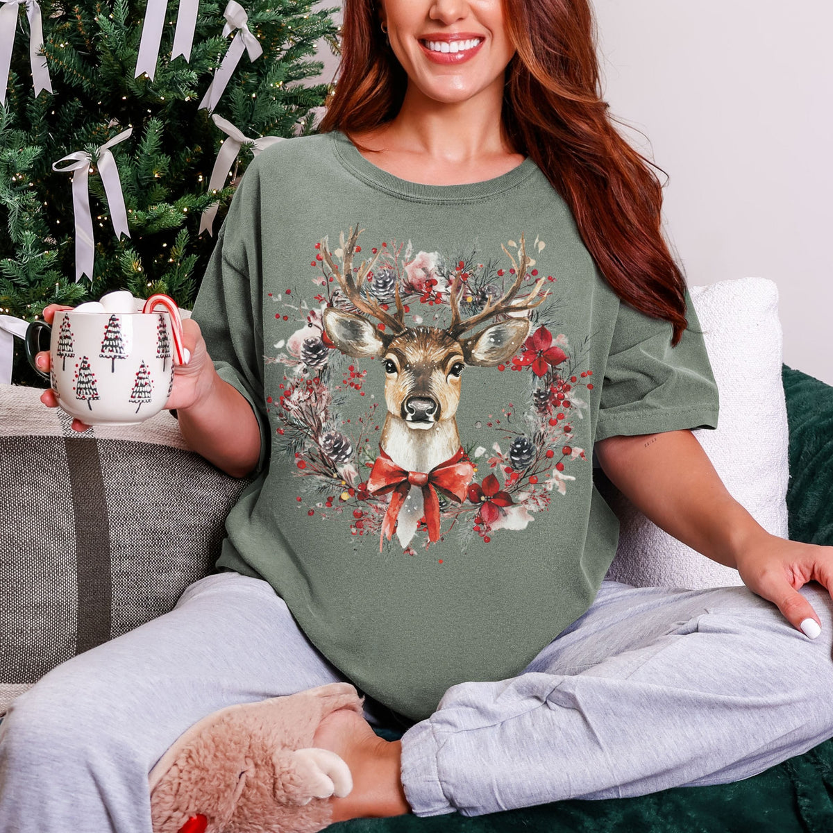 A woman sitting on a couch wearing an oversized sage green t-shirt with a festive Christmas deer graphic, holding a mug in front of a decorated Christmas tree.