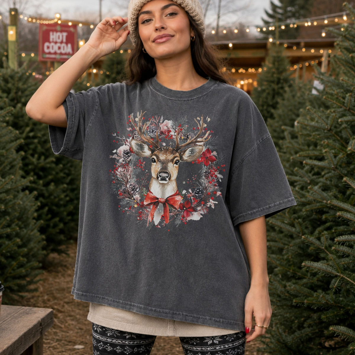 A woman wearing an oversized dark gray washed graphic t-shirt with a festive deer and wreath print, standing in a Christmas tree lot.