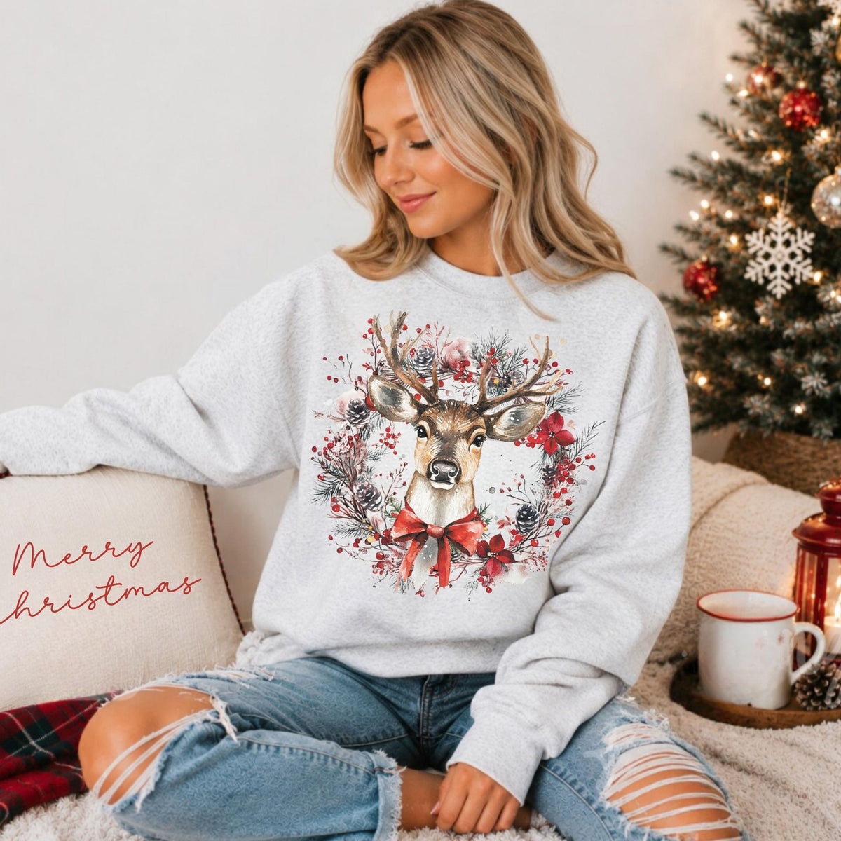 A woman sits on a couch wearing a light gray crewneck sweatshirt with a large watercolor deer and floral wreath graphic on the front. The scene is decorated for Christmas, with a lit tree, a mug, and a pillow that says 'Merry Christmas' in the background.