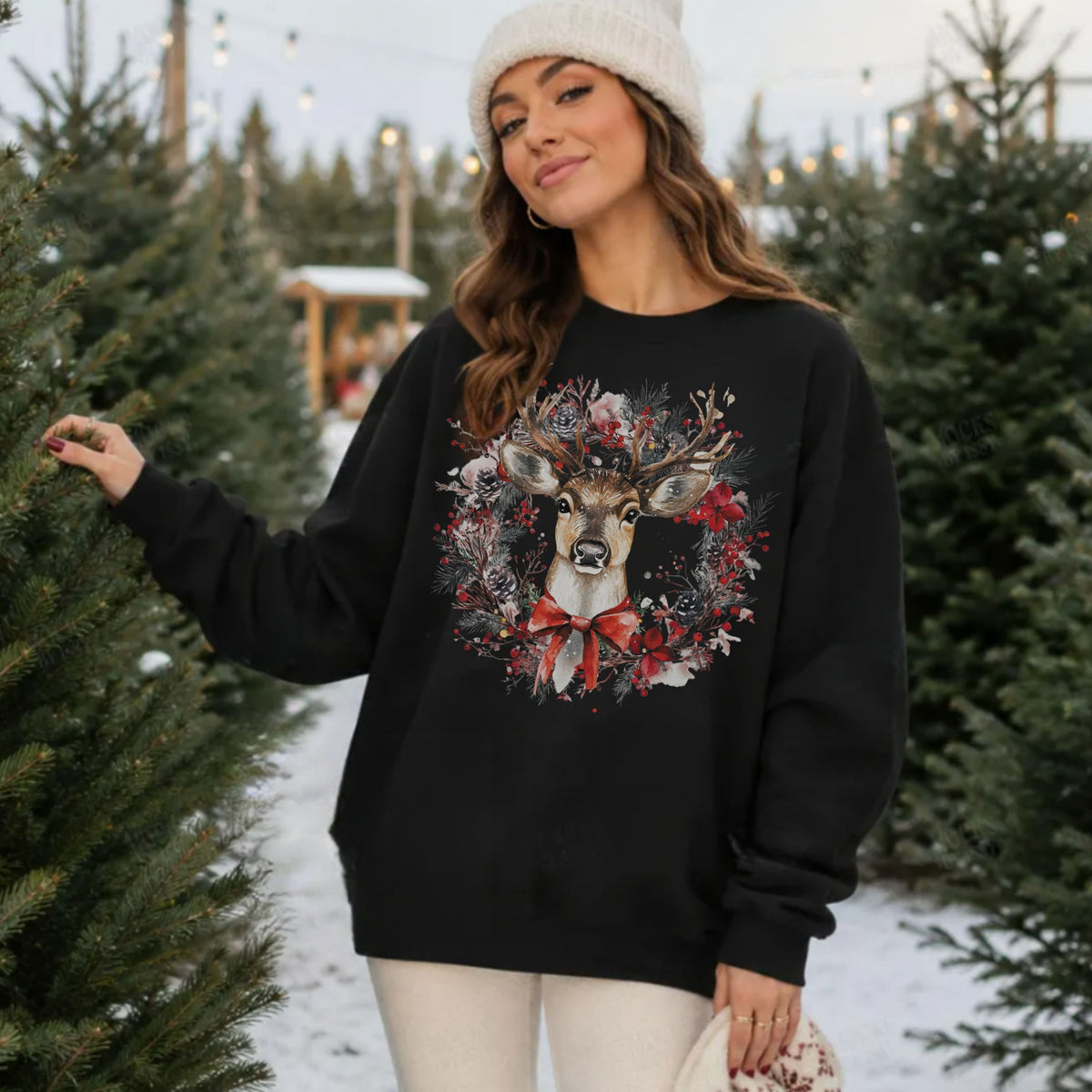 A woman wearing a black crewneck sweatshirt with a large festive deer wreath graphic, standing in a snowy Christmas tree lot.