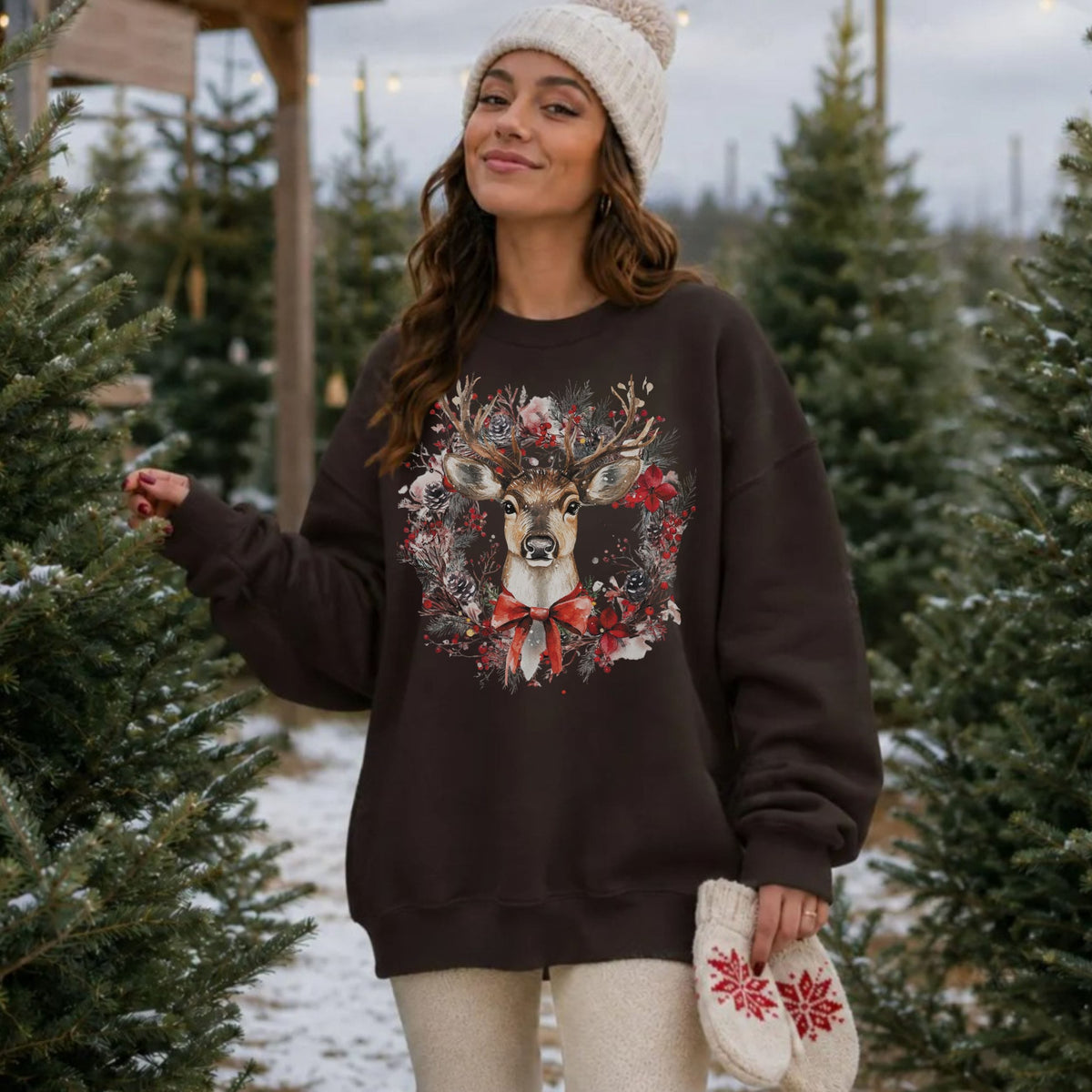 A woman wearing a dark brown crewneck sweatshirt with a festive deer wreath graphic, standing in a Christmas tree lot. She is also wearing a cream knit beanie and holding a matching cream knit mitten with red snowflake patterns.