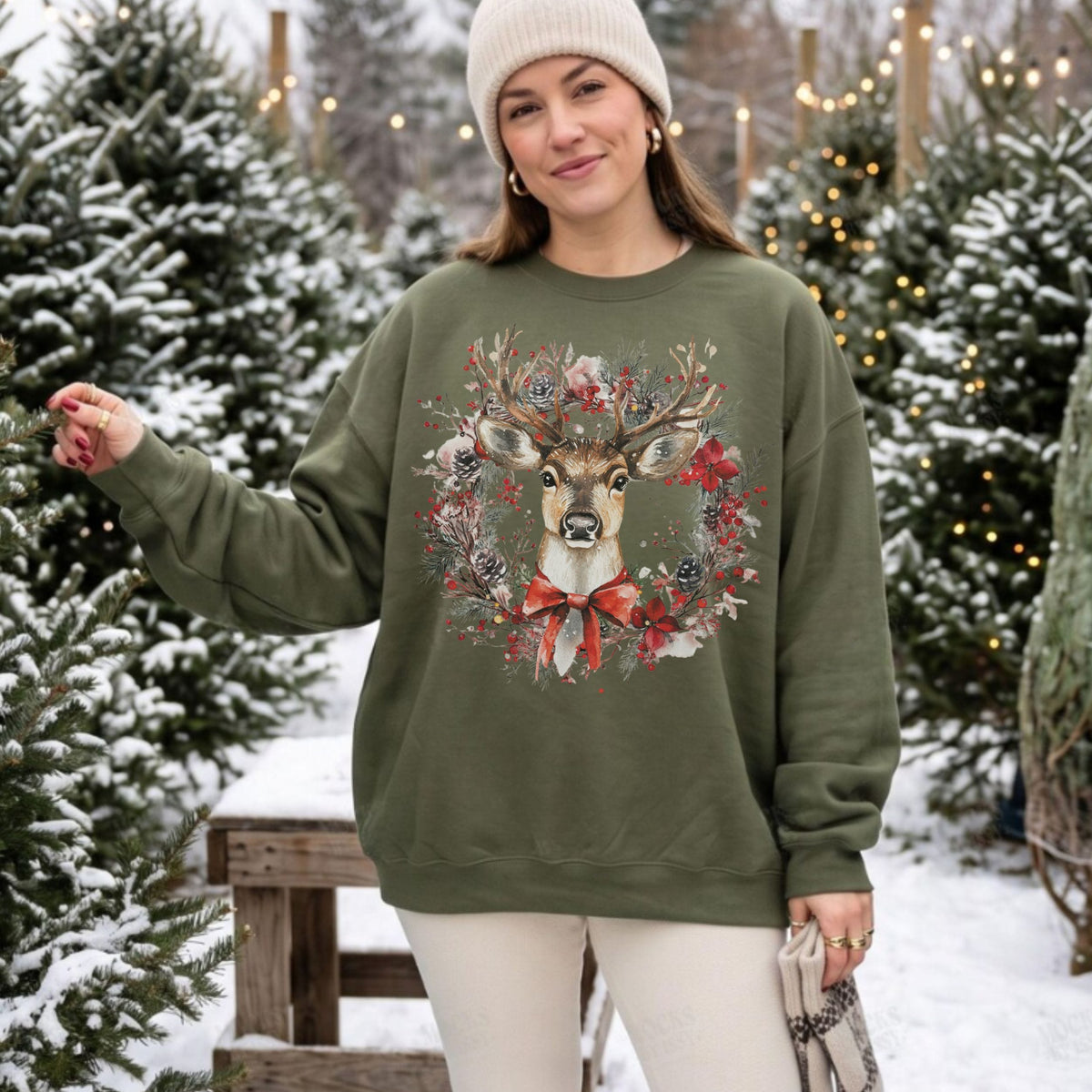 A woman wearing a military green crewneck sweatshirt with a detailed deer and wreath graphic, standing in a snowy outdoor setting with Christmas trees.