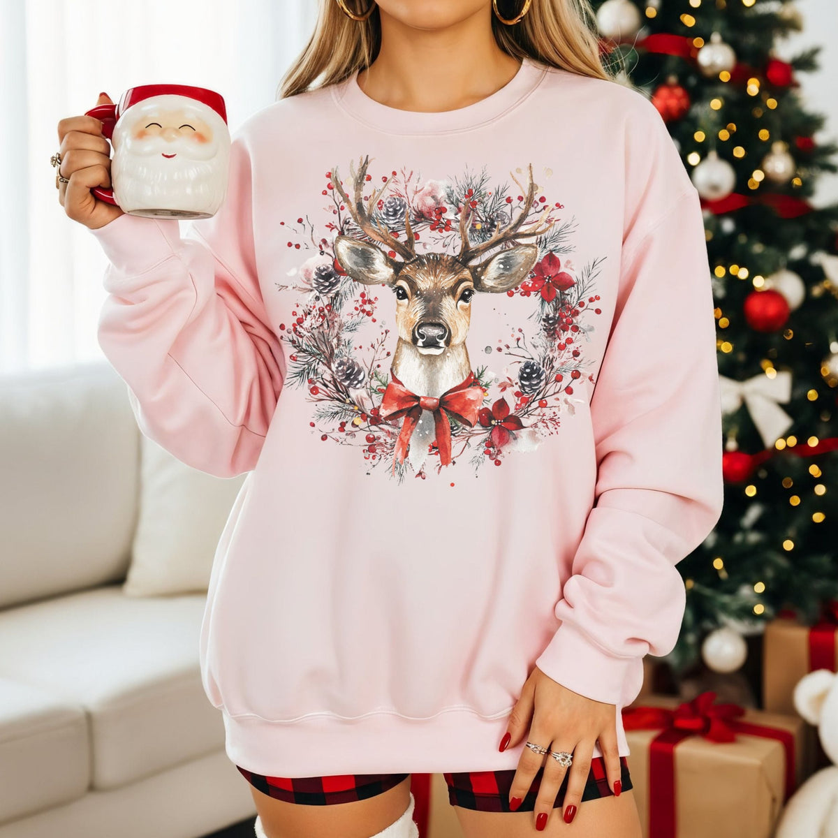 A woman wearing a light pink crewneck sweatshirt with a large festive graphic of a deer surrounded by a wreath and red bow, holding a Santa mug in a living room setting with a decorated Christmas tree in the background.