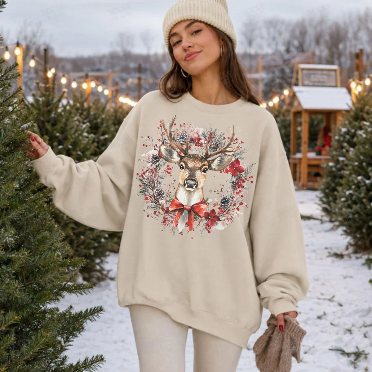 A woman wearing a cream-colored crewneck sweatshirt with a festive Christmas deer graphic, standing in a snowy outdoor Christmas tree lot.