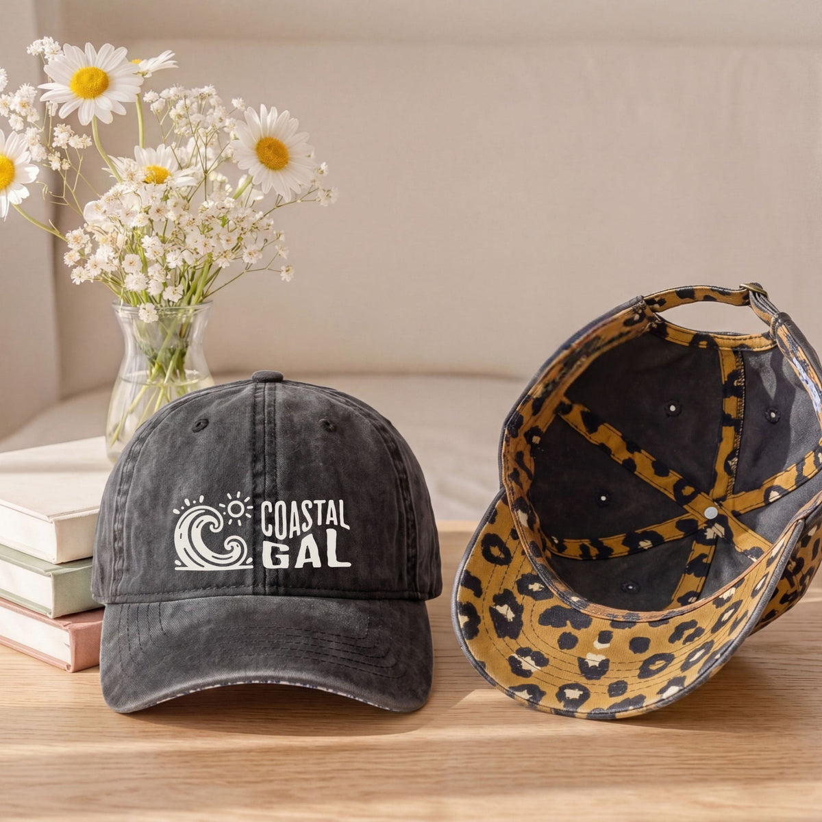 Two baseball caps displayed on a wooden table with a vase of daisies and books in the background. The cap on the left is washed black with a white 'COASTAL GAL' wave logo on the front.